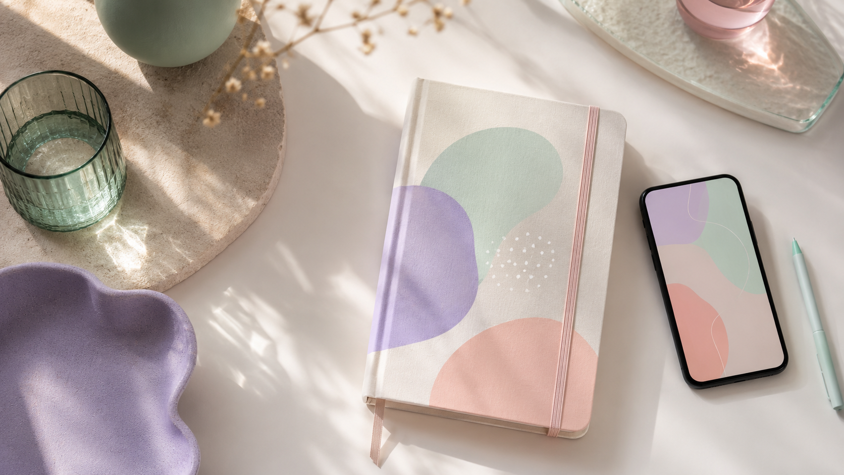 Soft flat-lay of a journal and gentle pastel shapes on a warm off-white surface